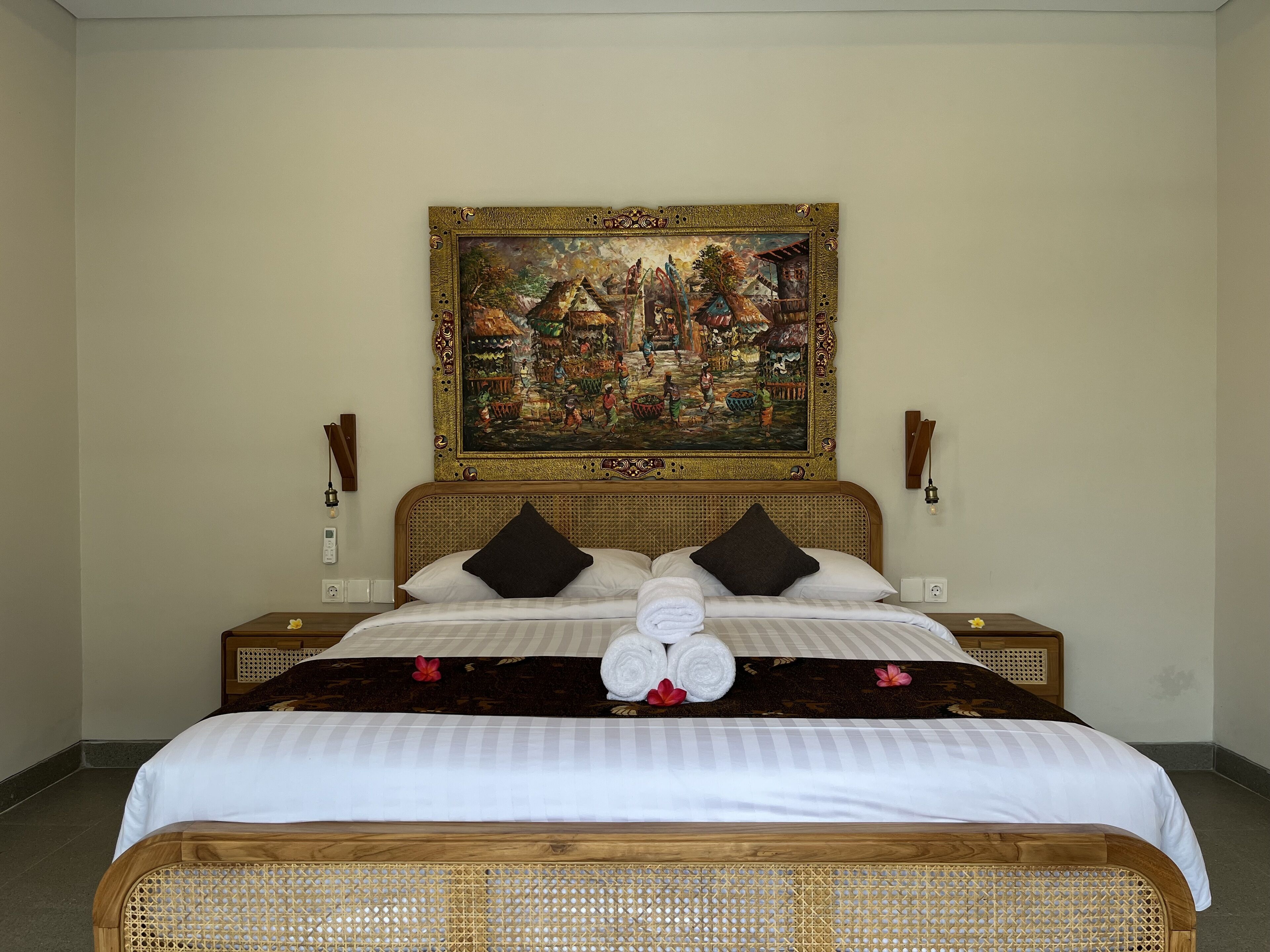 A neatly made bed with white linens, two black pillows, and a decorative pillow with a floral design. A painting of a village scene hangs above the bed.
