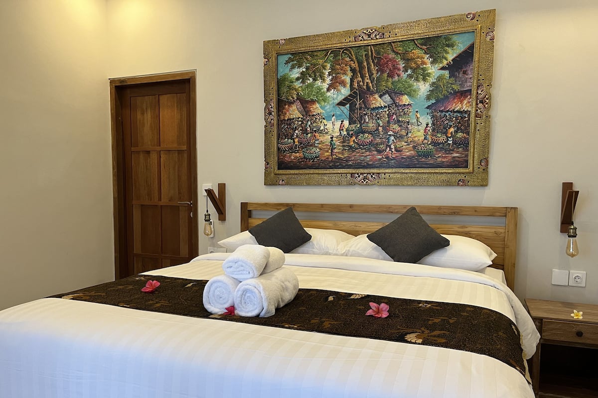 A neatly made bed with white linens and rolled towels, a wooden headboard, and a painting of a market scene above it.