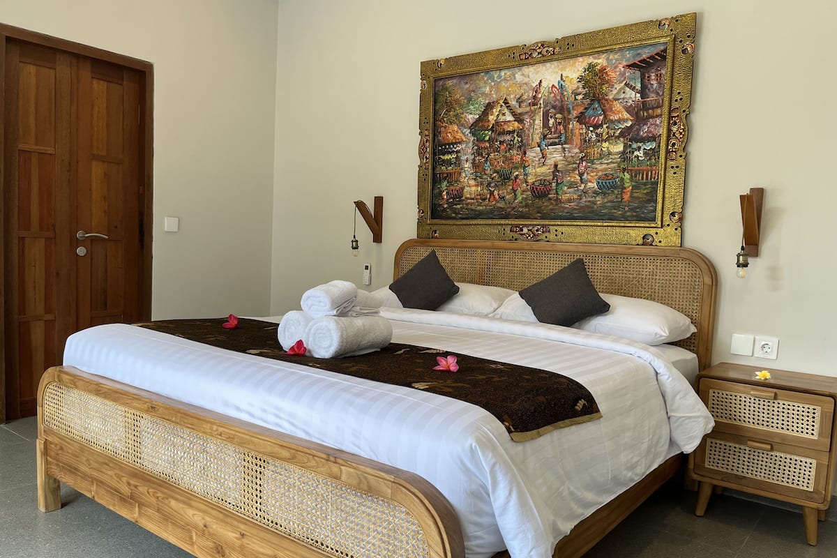 A neatly made bed with white linens, a rattan headboard, and a painting above it.