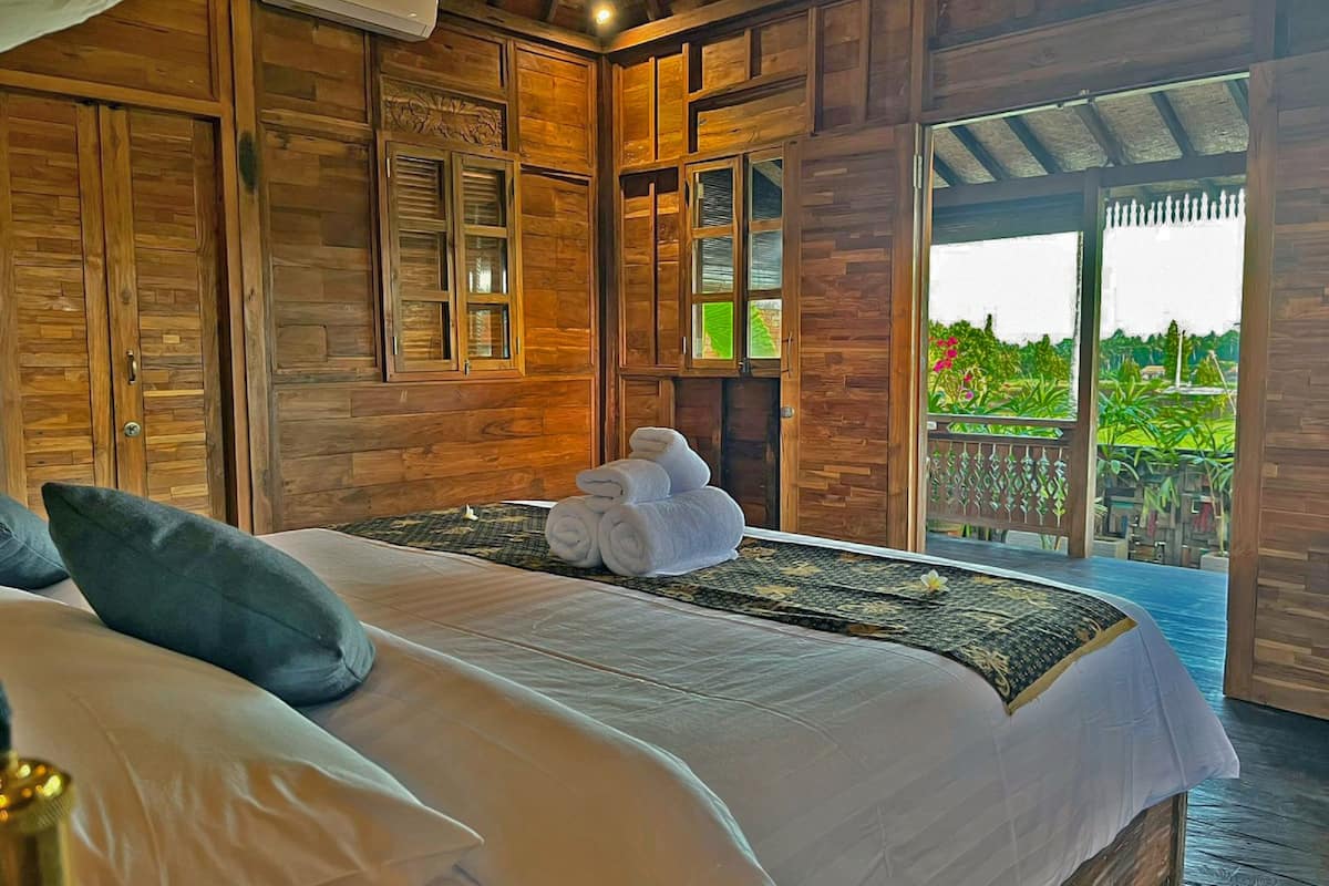 A bedroom with a large bed, wooden walls, and a balcony with a view of greenery.