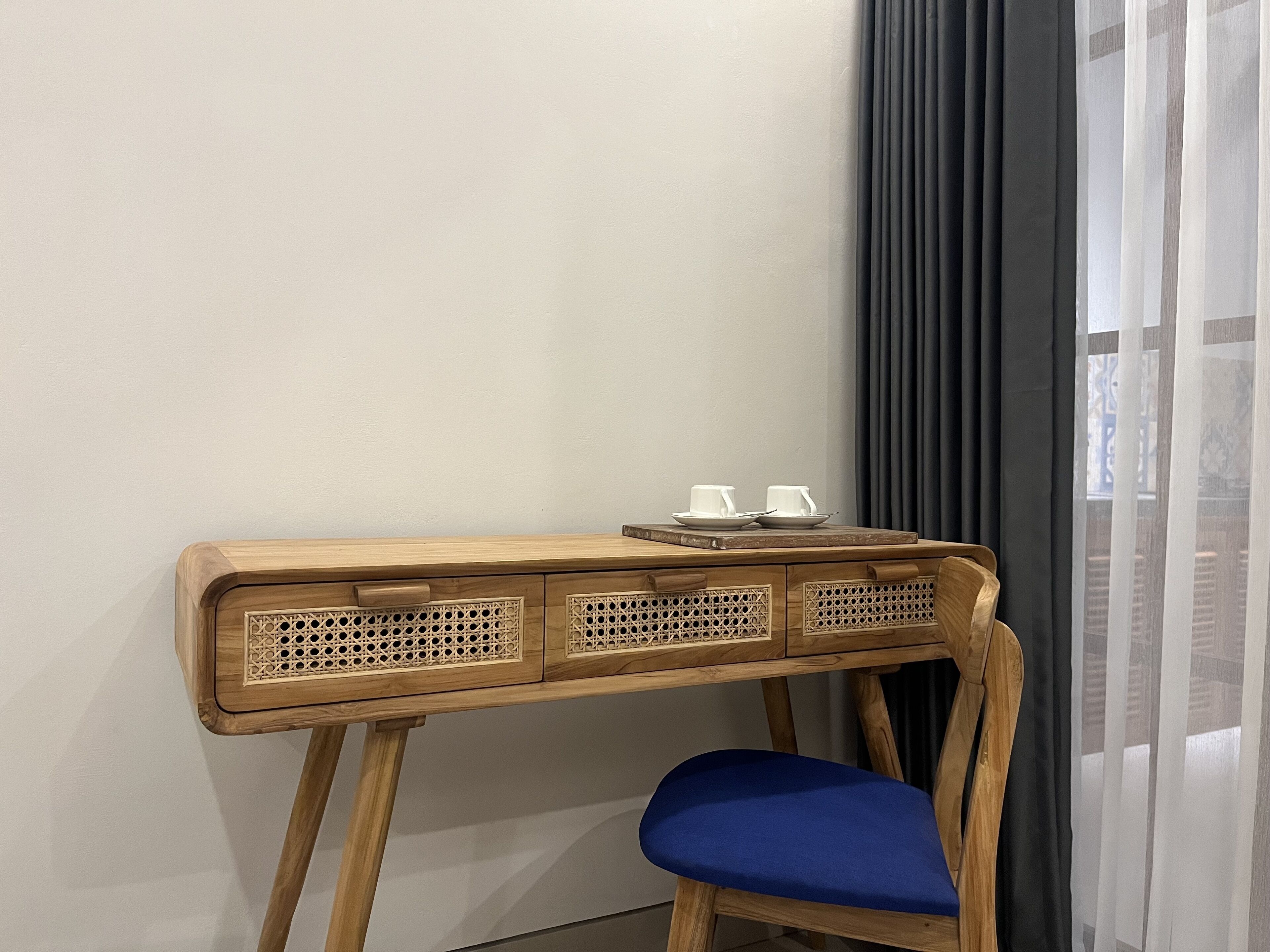 Un escritorio de madera con una silla azul, una mesita con dos tazas y platillos blancos, y una ventana con cortinas translúcidas.