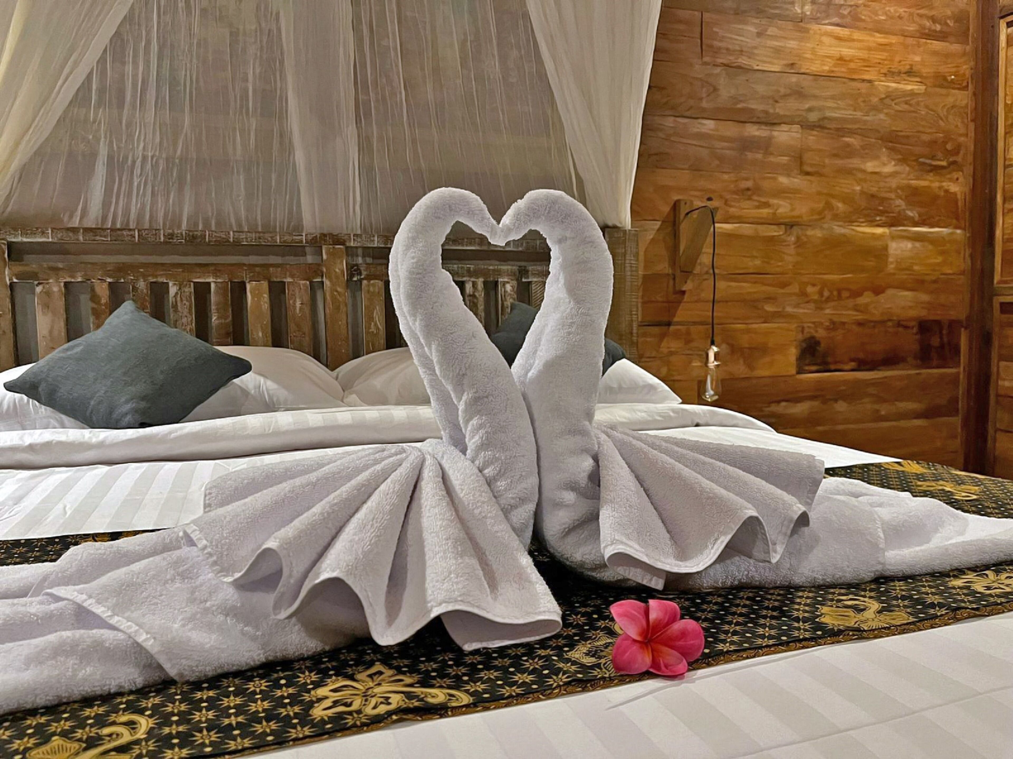 Una cama con ropa blanca, un arreglo de toallas en forma de corazón y un cojín decorativo.