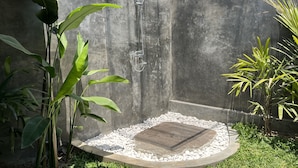 Un área de ducha de hormigón con una pérgola de madera, rodeada de exuberante vegetación y guijarros blancos.