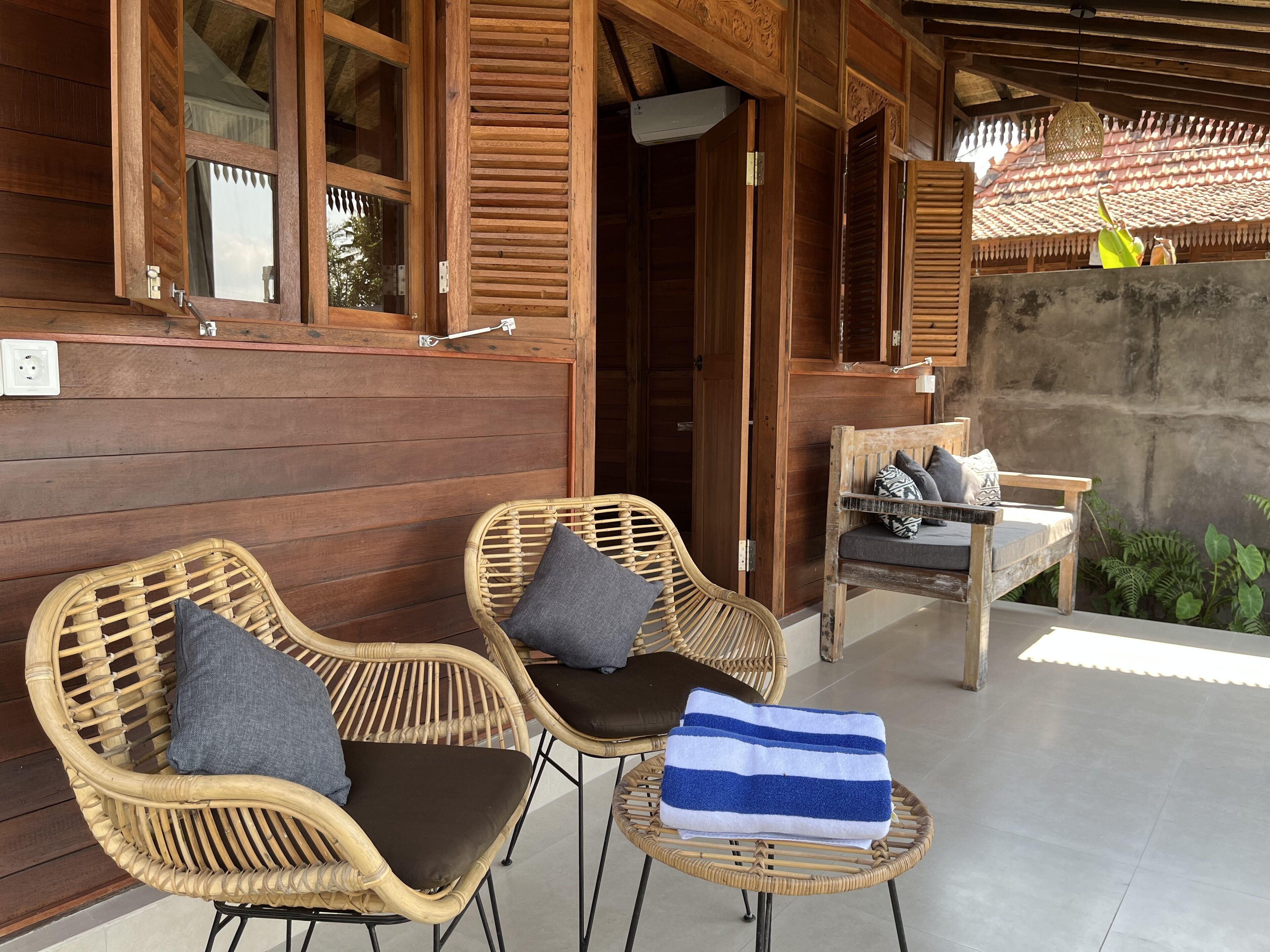 Exterior de madera con dos sillas de mimbre y un banco, una toalla sobre el banco, y una pared de concreto con vegetación.