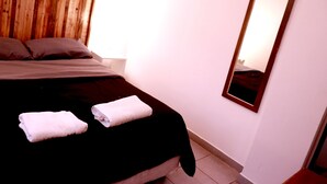 A bedroom with a wooden headboard, a bed with a black and grey bedspread, two white towels, a nightstand, and a mirror on the wall. - Ruda Hostel (Buenos Aires)
