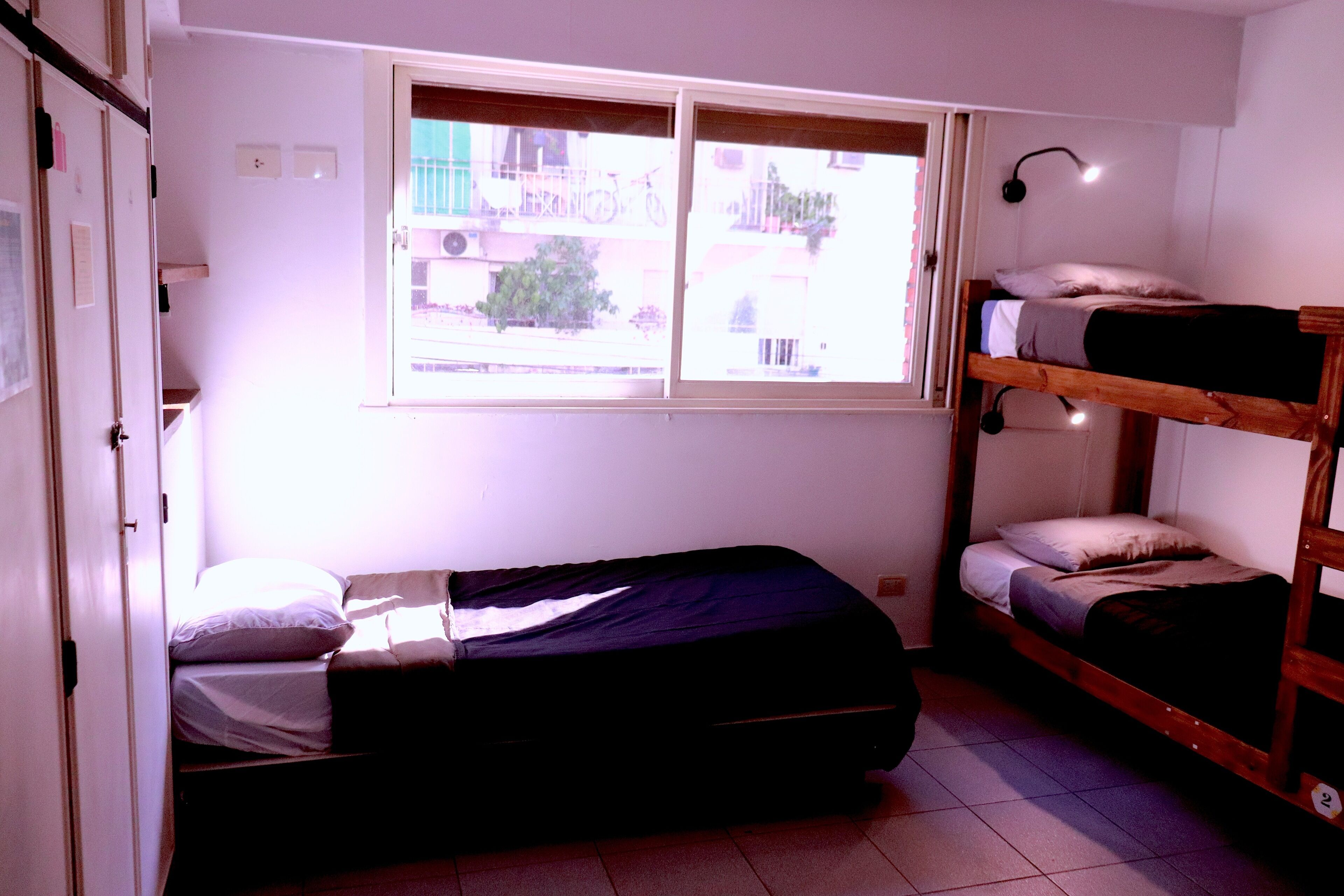 A dormitory room with a bunk bed, a window with a view of the outside, and a bedside lamp.