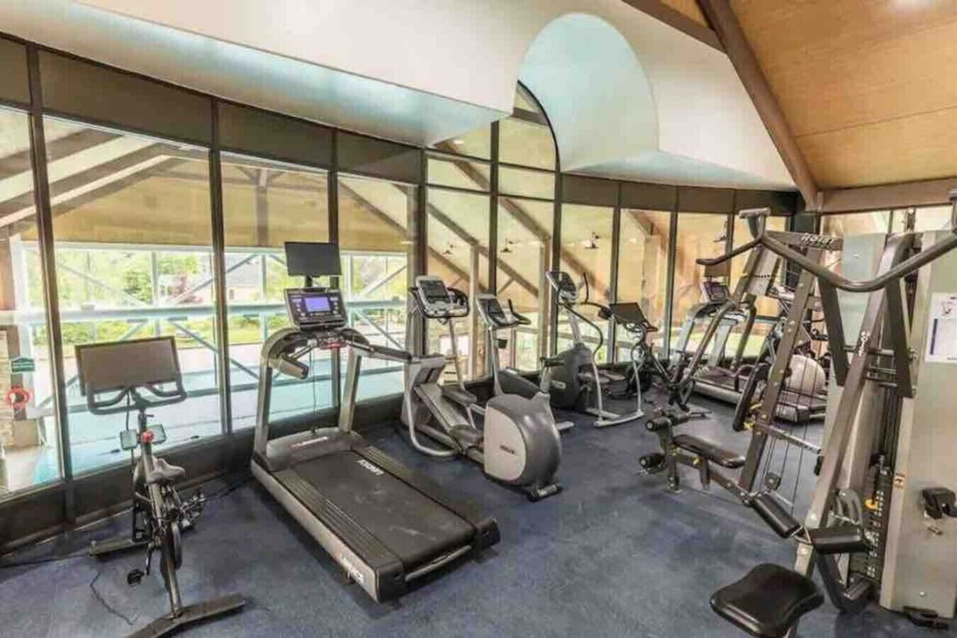 Un gimnasio con máquinas de cardio, equipo de pesas y grandes ventanales que ofrecen vistas al exterior.