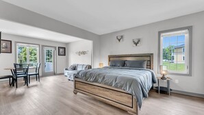 A spacious bedroom with a large bed, a desk, and a chair, featuring a view of the outdoors through large windows.