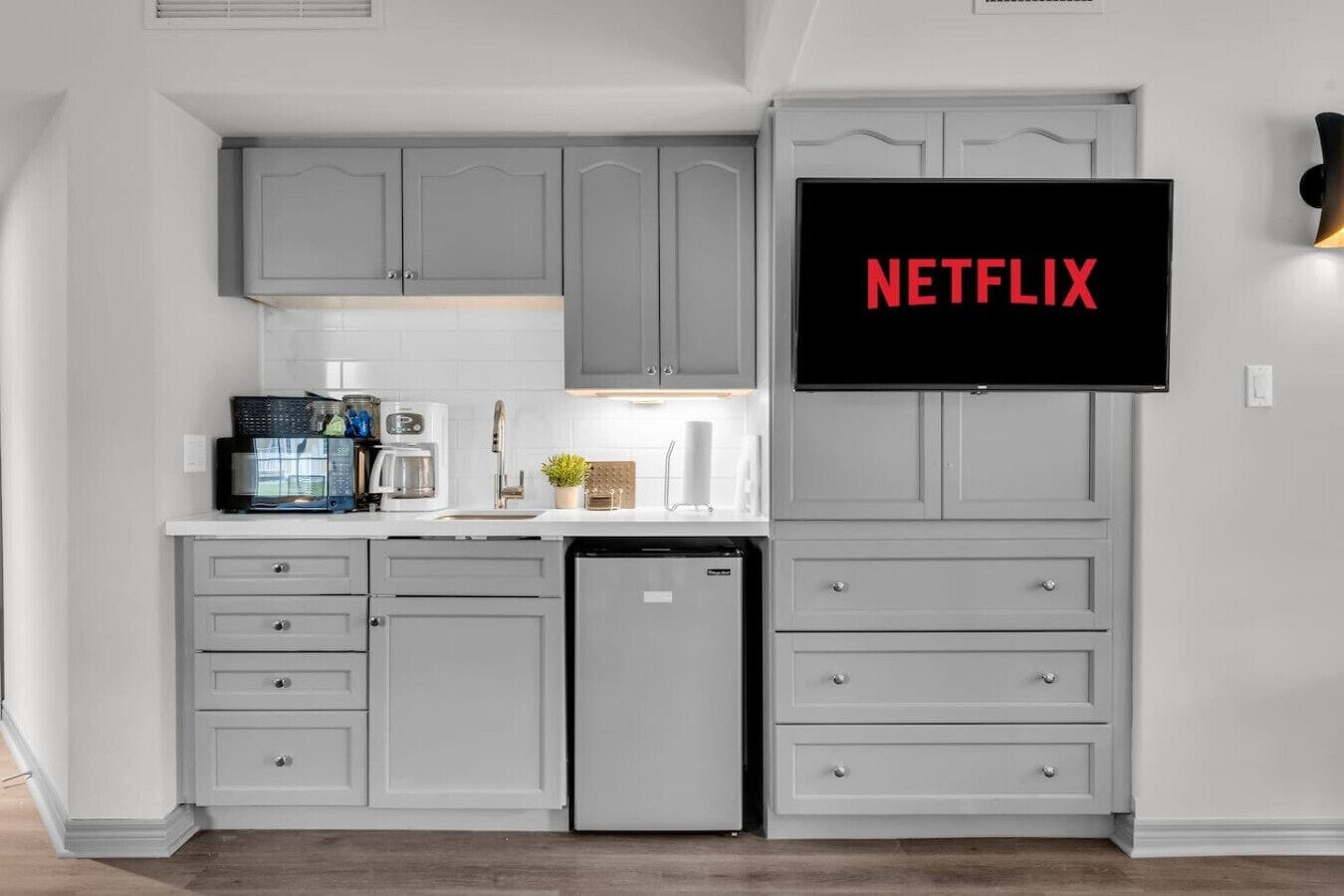 A modern kitchen with grey cabinetry, a built-in dishwasher, and a flat-screen TV mounted above the counter displaying the Netflix logo.