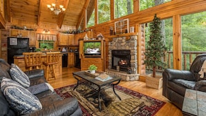 A cozy cabin interior with a stone fireplace, wooden furniture, and large windows overlooking a forest. - Happily Ever After (Gatlinburg)