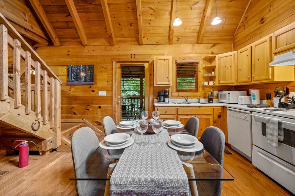 A wooden cabin interior with a dining table set for six, kitchen area with appliances, and a large window with blinds.