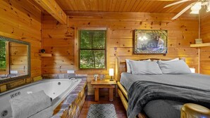 A wooden cabin room with a large bed, a wooden headboard, a small table with a lamp, a window with blinds, and a framed picture on the wall. - Sunrise Woods Retreat (Sevierville)