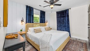 Un dormitorio con cama, mesita de noche, ventilador de techo y ventana con cortinas.