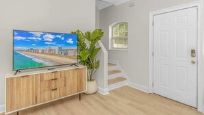 A room with a wooden cabinet, a television displaying a beach scene, a large plant, a white door, and a staircase with white walls and wooden steps. - 4-min Walk to Ocean|2 King Suites|Beach Retreat (Myrtle Beach)