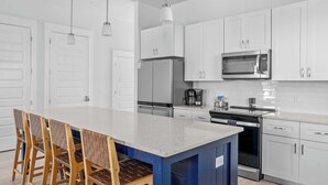 A modern kitchen with white cabinets, a stainless steel refrigerator, a microwave, and a built-in oven. There is a large island with a blue base and a white countertop. - 6-Min Walk to Beach | Heated Pool | Fenced Yard (Panama City)