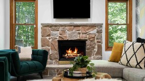 A cozy living room with a stone fireplace, green and grey sofas, and a flat-screen TV mounted above the fireplace. - 5BR|Private Lake|Pools|Hot Tub|Near Camelback (East Stroudsburg)