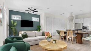 Un salon moderne avec un canapé, des fauteuils verts, une table basse en bois et un coin repas comprenant des chaises, avec une cuisine en arrière-plan.