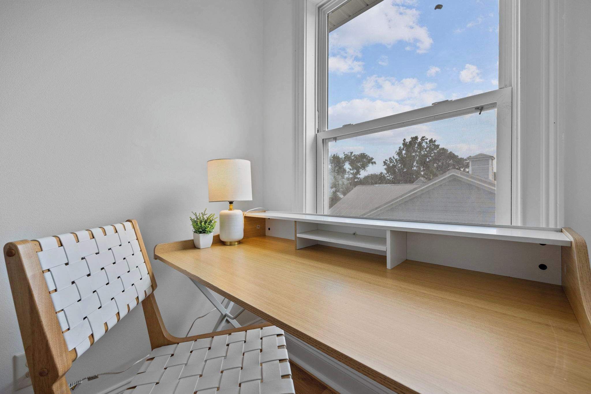 A corner desk with a white chair, a small table with a lamp, and a window with a view of trees and a house.