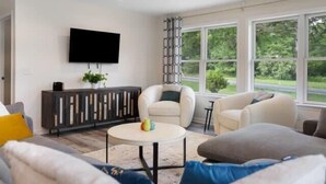Ein modernes Wohnzimmer mit einem an der Wand befestigten Flachbildfernseher, einem runden Couchtisch mit bunten Objekten und einem großen Fenster mit Blick auf Grünfläche.