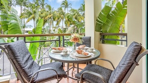 A balcony with a round table set for two, surrounded by wicker chairs, overlooking a tropical landscape with palm trees and a clear sky. - Unique Tropic Escape 111 (Cairns North)