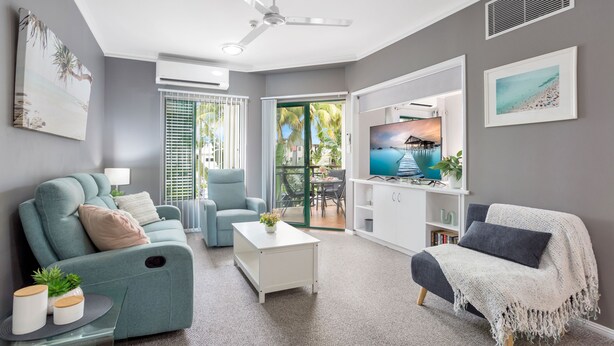 A modern living room with a sofa, armchair, and coffee table. There is a TV on a stand, a ceiling fan, and a sliding glass door leading to an outdoor area. - Unique Tropic Escape 111 (Cairns North)