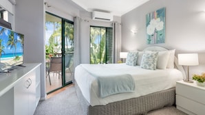 A modern bedroom with a bed, bedside tables, a TV, and a view of the beach through a sliding door. - Unique Tropic Escape 111 (Cairns North)