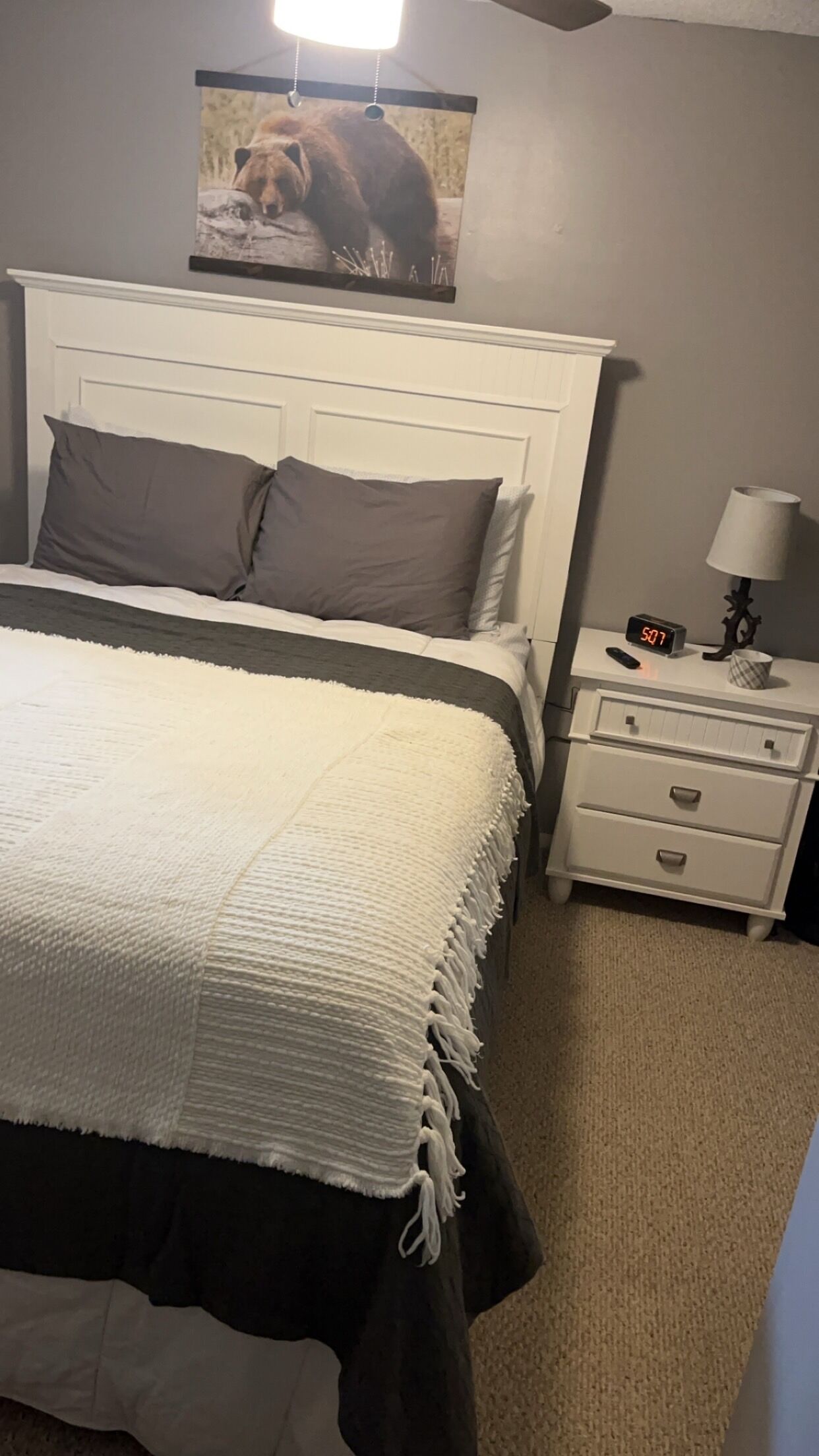 Une chambre avec un lit blanc, des oreillers gris, une table de chevet blanche avec une lampe, et un tableau représentant un ours accroché au mur.
