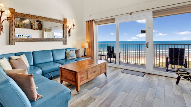 A spacious living room with a blue sofa, wooden coffee table, and a large mirror. It features a sliding glass door leading to a balcony with a view of the ocean. - Penthouse Dahktari is a true beach front high-rise (10th floor) condominium (South Padre Island)