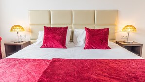 Un lit bien fait, recouvert d’une couvre-lit rouge et blanche, avec deux oreillers rouges et une tête de lit beige. Deux lampes de chevet sont posées sur des tables de chevet en bois.