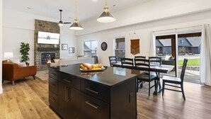 A spacious living area with a stone fireplace, a dining table with chairs, and a large island with a black countertop.