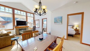A spacious living area with a wooden dining table, comfortable seating, a fireplace, and large windows offering a view of the outdoors. - Settler's Crossing #42 (Sun Peaks)