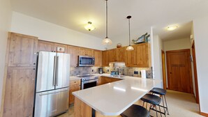 A modern kitchen with wooden cabinets, stainless steel appliances, a large island with bar stools, and pendant lighting. - Settler's Crossing #42 (Sun Peaks)