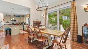 A spacious kitchen with wooden flooring, a dining area with a large wooden table and chairs, and a view of greenery through large windows. - Off Belay | Luxury Creekside Home, Screened Porch, Fireplace & Gourmet Kitchen (Chincoteague Island)