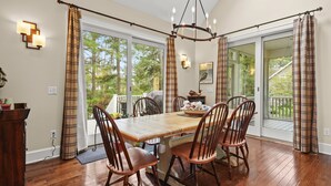 A dining area with a wooden table and chairs, a chandelier, and a view through large sliding glass doors to an outdoor deck. - Off Belay | Luxury Creekside Home, Screened Porch, Fireplace & Gourmet Kitchen (Chincoteague Island)