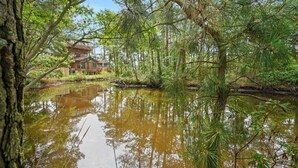 A house surrounded by a forest with a pond in the foreground. - Off Belay | Luxury Creekside Home, Screened Porch, Fireplace & Gourmet Kitchen (Chincoteague Island)