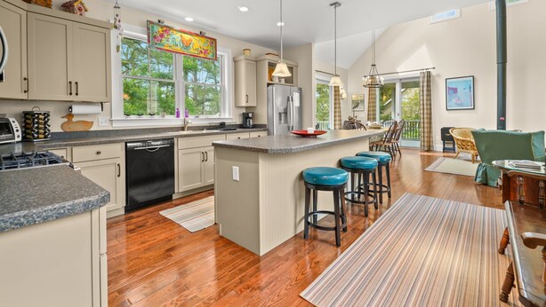 A spacious kitchen with wooden flooring, a central island with bar stools, and a dining area with a table and chairs. - Off Belay | Luxury Creekside Home, Screened Porch, Fireplace & Gourmet Kitchen (Chincoteague Island)