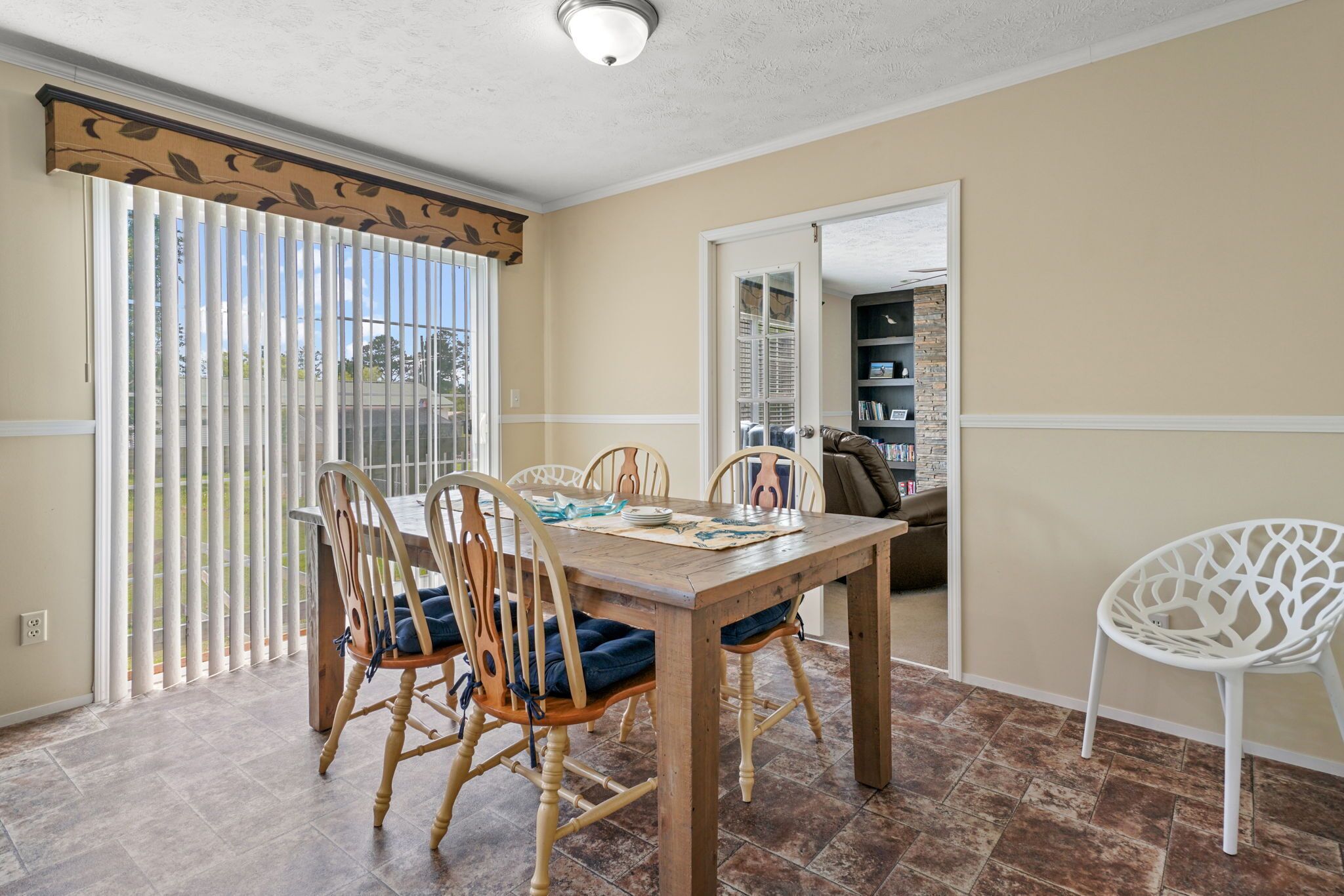 A dining area with a wooden table and chairs, a white chair, and a view through a sliding glass door.