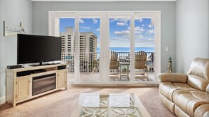 A living room with a TV, a leather sofa, and a balcony with a view of buildings and the sea.
