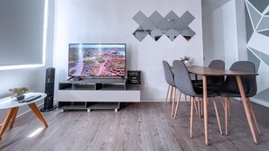 Uma sala de estar moderna com uma televisão de tela plana, uma mesa de jantar com cadeiras, uma pequena mesa lateral e um espelho decorativo na parede.