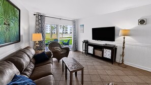 A living room with a leather sofa, a wooden coffee table, a TV mounted on the wall, and a view of the outdoors through large windows.