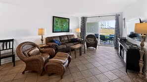 A living room with a brown leather sofa, wicker chairs, a wooden coffee table, and a view of the beach through the glass door.