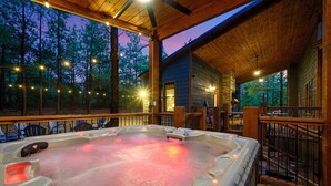 A hot tub on a wooden deck with string lights, surrounded by a forest at dusk. - Star-Spangled Fun | Arcade + Outdoor Oasis + Putt-Putt + EV Outlet (Broken Bow)