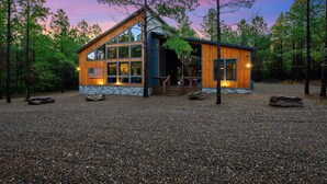A modern log cabin with large windows, a stone foundation, and a gravel driveway surrounded by trees. - Star-Spangled Fun | Arcade + Outdoor Oasis + Putt-Putt + EV Outlet (Broken Bow)
