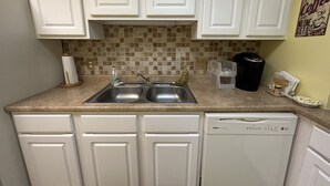 Una cocina con armarios blancos, un lavamanos doble, una cafetera y un escurreplatos.