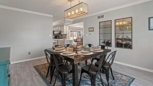 Un coin repas avec une table en bois dressée pour six personnes, entourée de chaises sombres. Un lustre moderne est suspendu au-dessus.