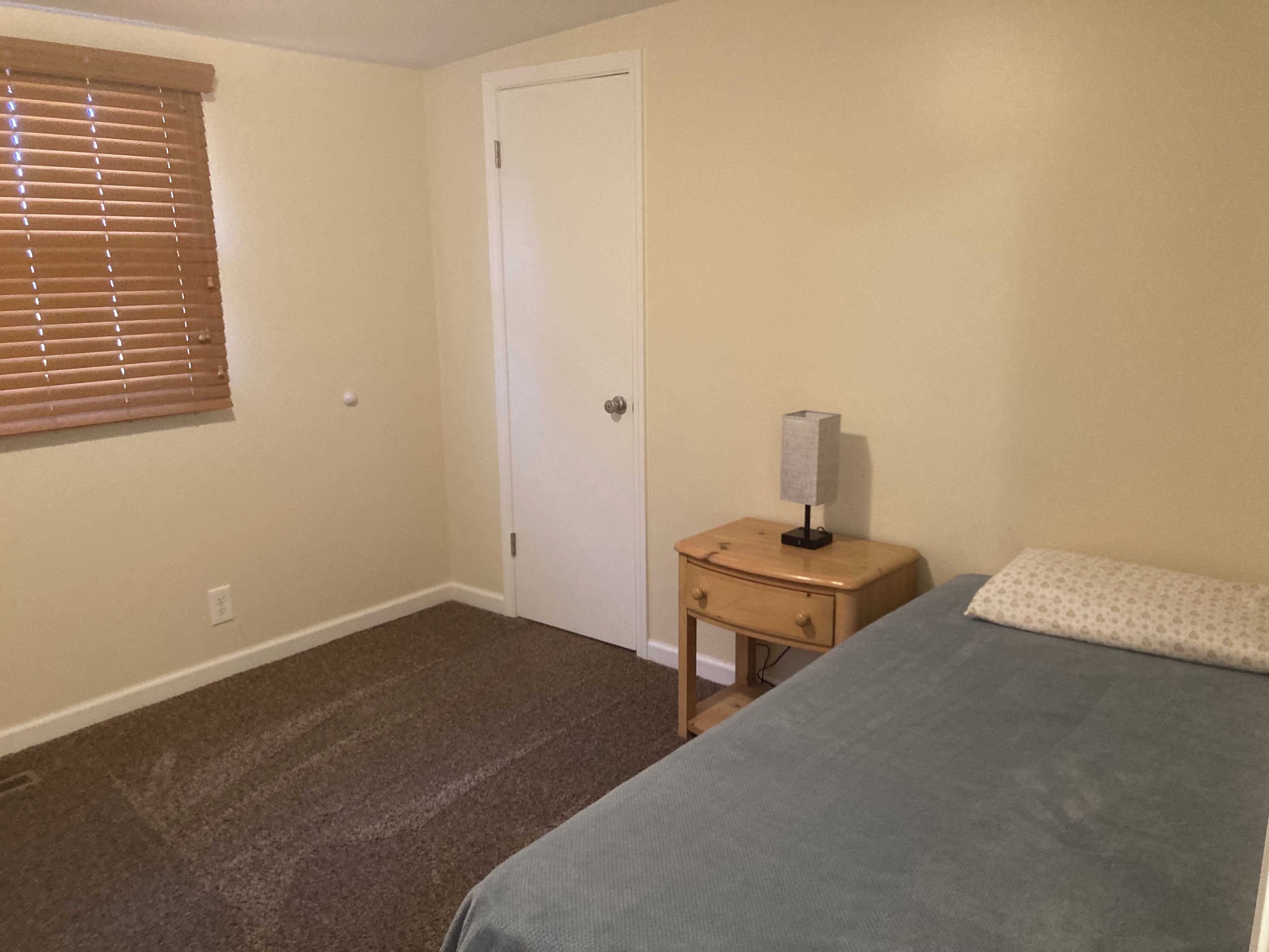 A bedroom with a single bed, a nightstand with a lamp, a door, and a window with blinds.