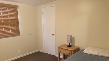 A bedroom with a single bed, a nightstand with a lamp, a door, and a window with blinds.