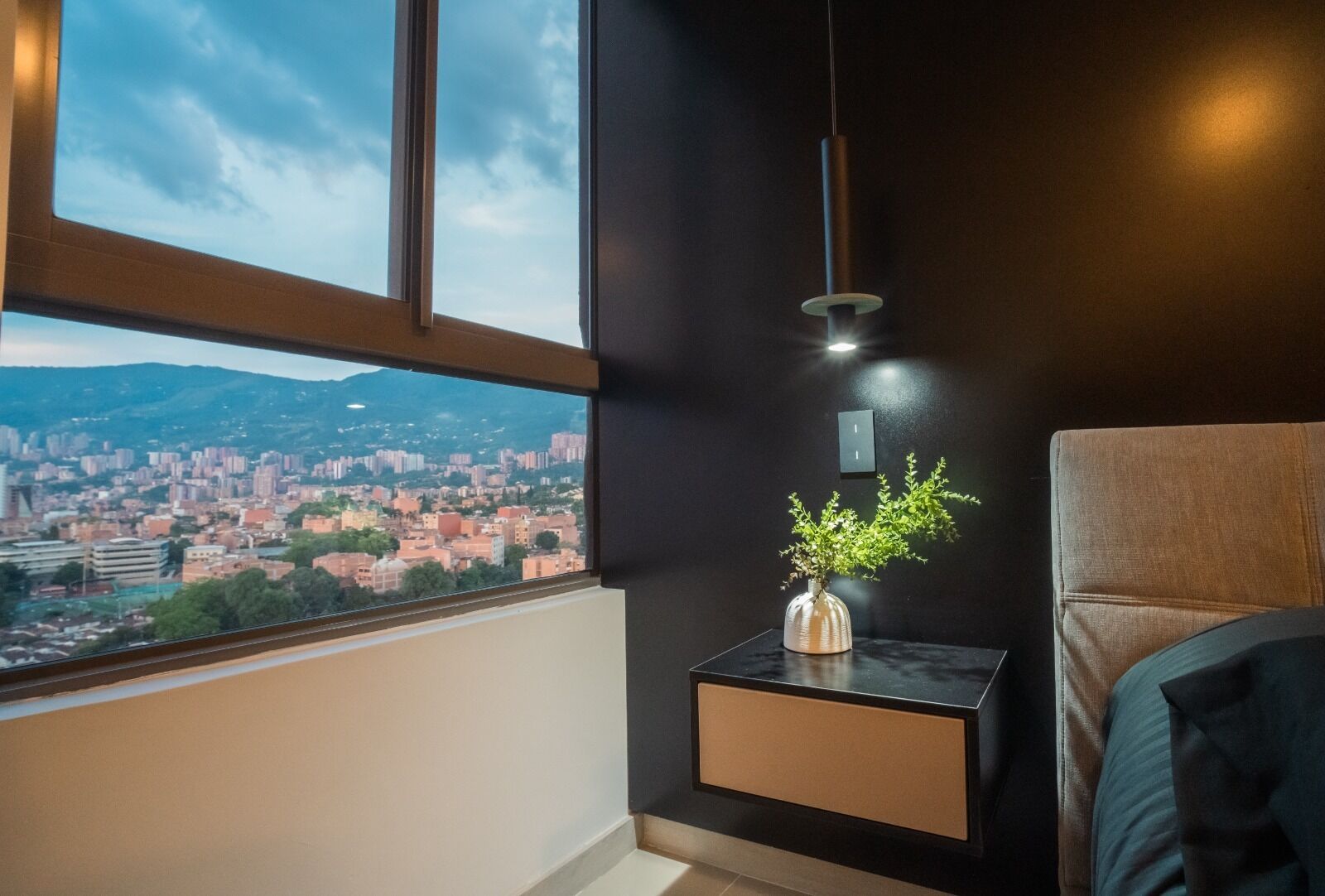 A modern room with a large window overlooking a cityscape, a small table with a plant, and a hanging light fixture. - Apartment Luxe with view of Poblado (Envigado)