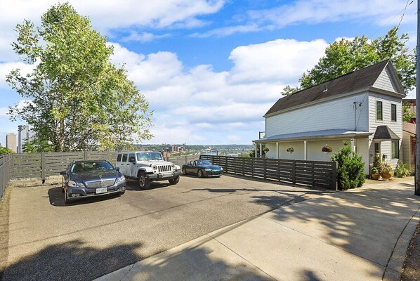 A residential driveway with three cars parked, a white house with a gabled roof, and a large tree in the background. - City View Sanctuary ~ Hot Tub, Abundant Parking, 3 Decks, Fenced Yard, Fire Pit (Pittsburgh)