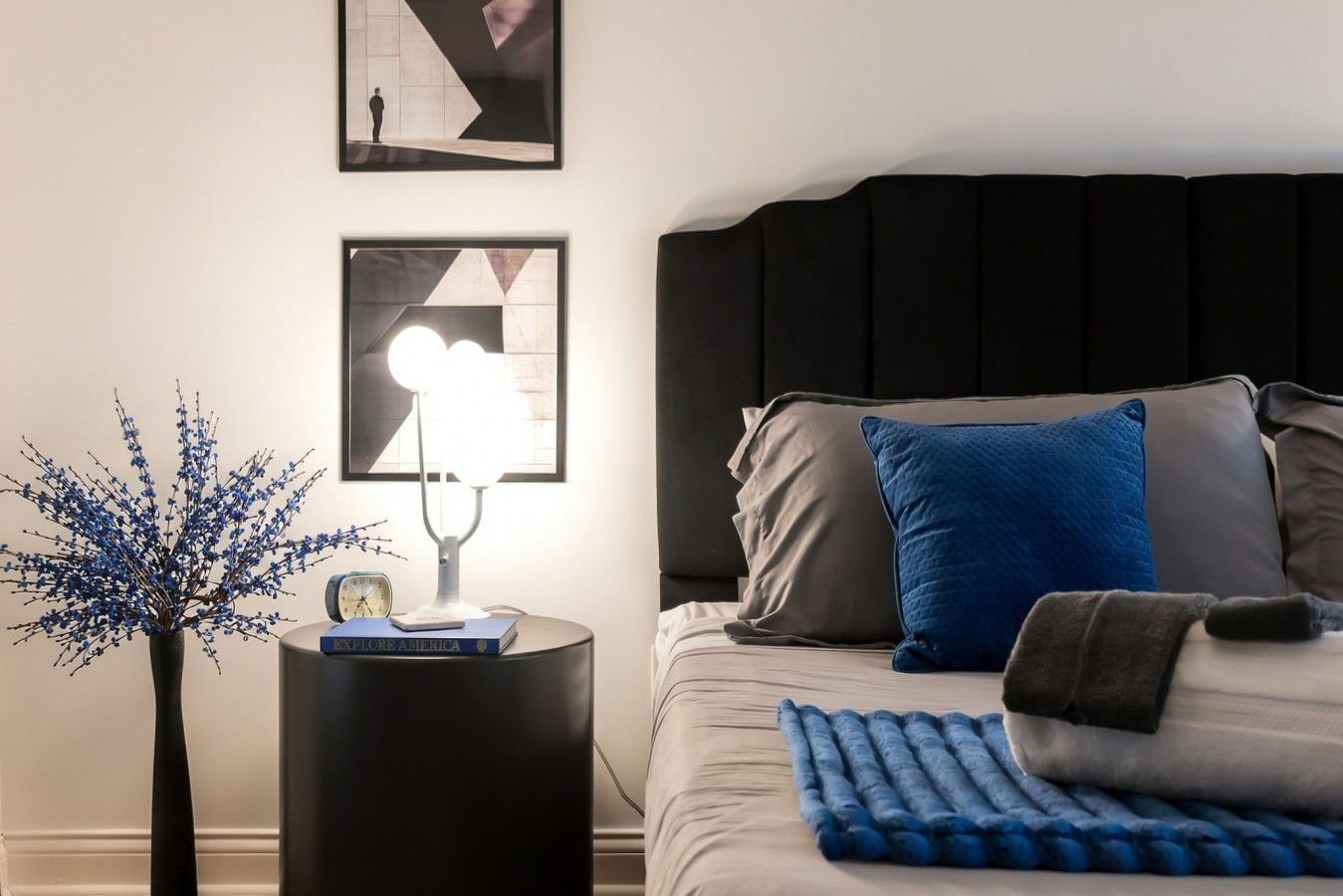 A bedroom with a bed, a bedside table with a lamp, a clock, and a vase with blue flowers.